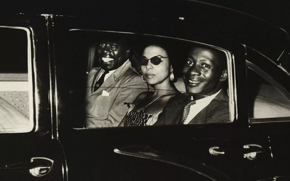 Black and white photo of three people sat in a car taken from outside the window.