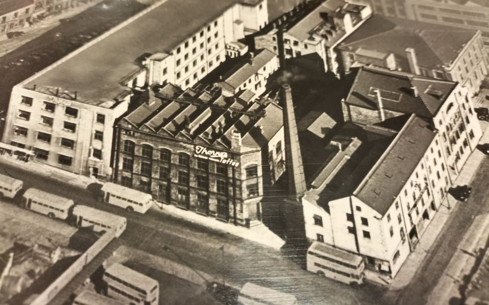 A black and white photo of the Thorne's cocoa works near Leeds Grand Theatre with a chimney billowing smoke, a sign reading Thorne's Toffee and buses all around