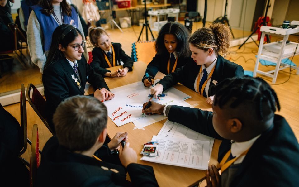 School children sitting round a table writing words on a large sheet of paper.