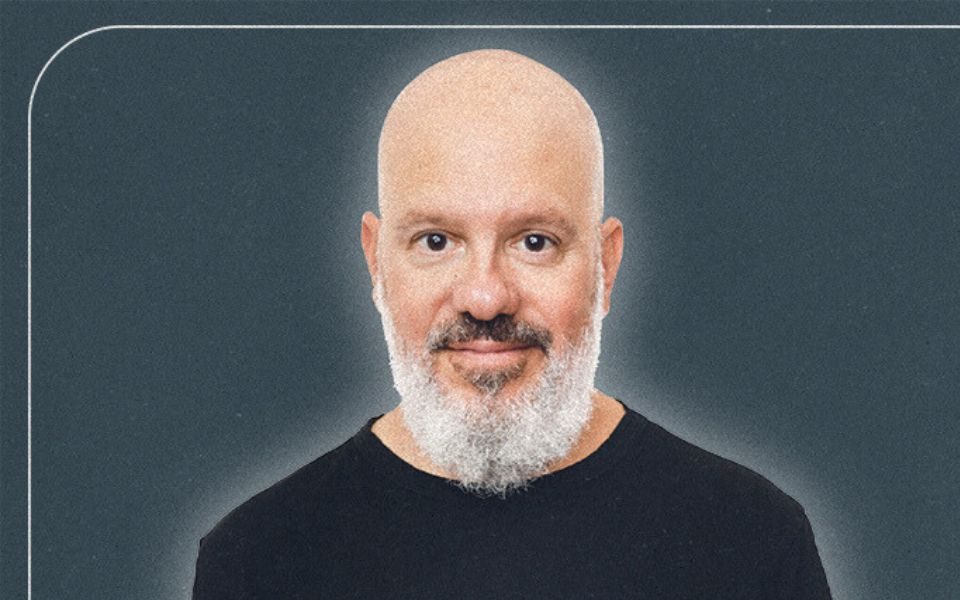 David Cross from the shoulders up with a neutral expression wearing a black t-shirt stood against a charcoal background.