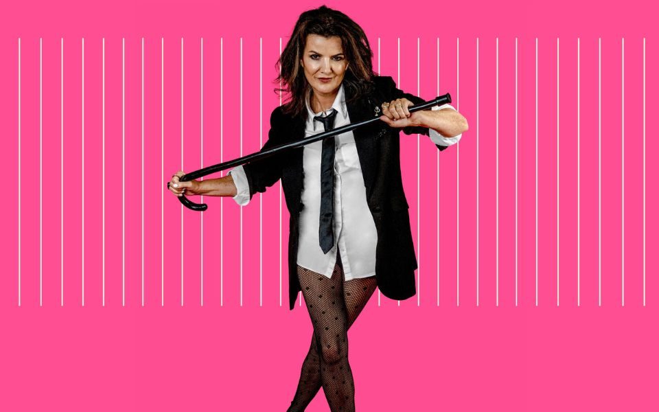 Deirdre O'Kane in a suit jacket, black tie, white shirt and just semi-sheer tights holding a cane across her chest with both hands stood against a pink background.