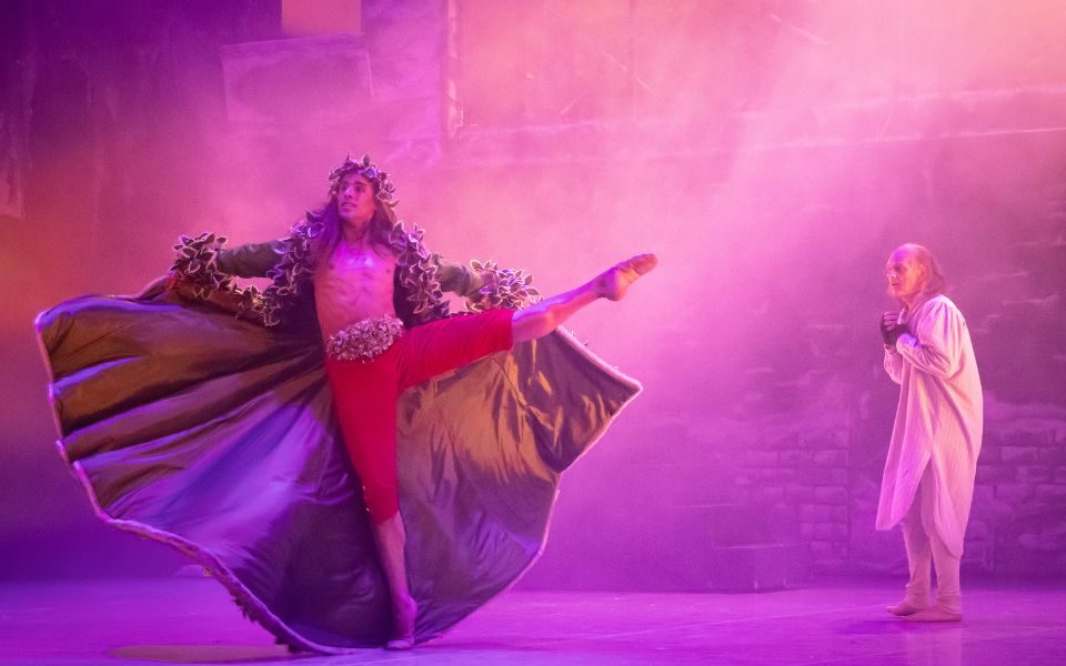 A dancer with their leg outstretched dressed in red trousers with a big cloak behind them and adorned in ivy leaves. A dancer dressed as Scrooge in a white nightdown stands in the background.