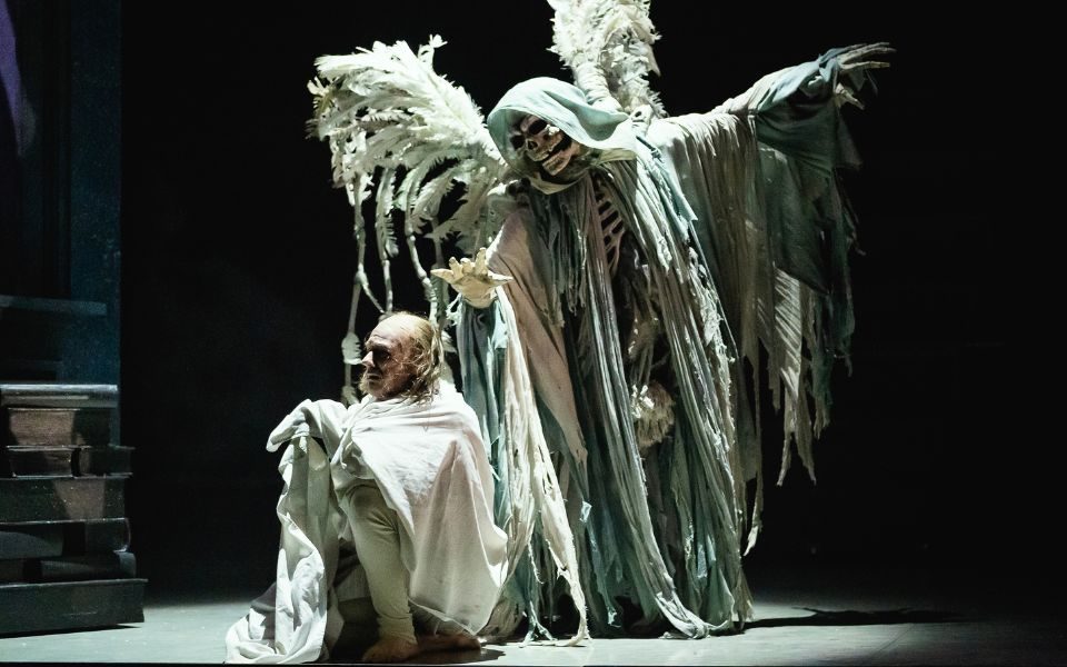 A dancer dressed in heavy layers of cloaks and a skull mask with large grey wings looms over a dancer dressed in a white nightgown as Scrooge.