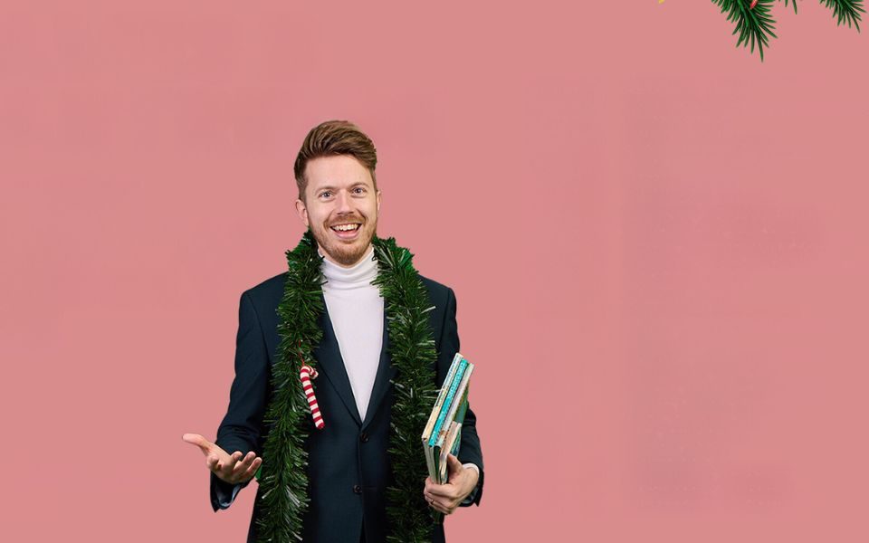 James B Partridge in a navy blazer and white turtle neck with some green tinsel round his neck that has a candy cane attached to it. He is holding some books. A little bit of Christmas tree appears in the top right hand corner.