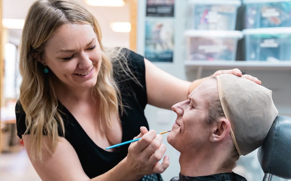 Harriet Rogers smiling as she paints Jonathan Hanks's face. He wears a bald cap as she holds his head.