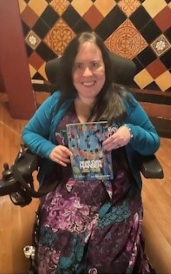 Lauren Fordham in her wheelchair smiling at the camera and holding up a programme for Dear Evan Hansen