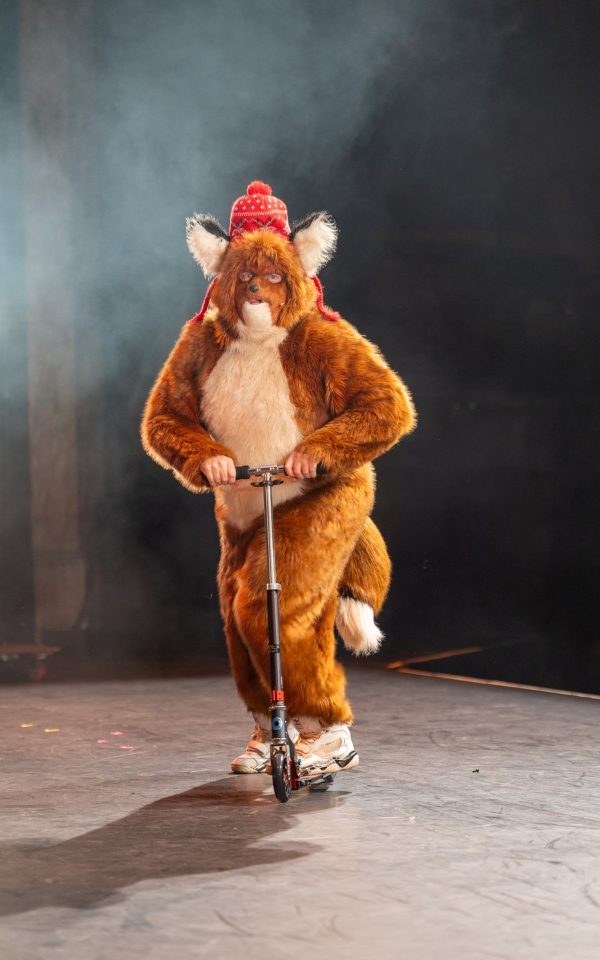 Leigh Francis as The Urban Fox, riding a scooter on stage.