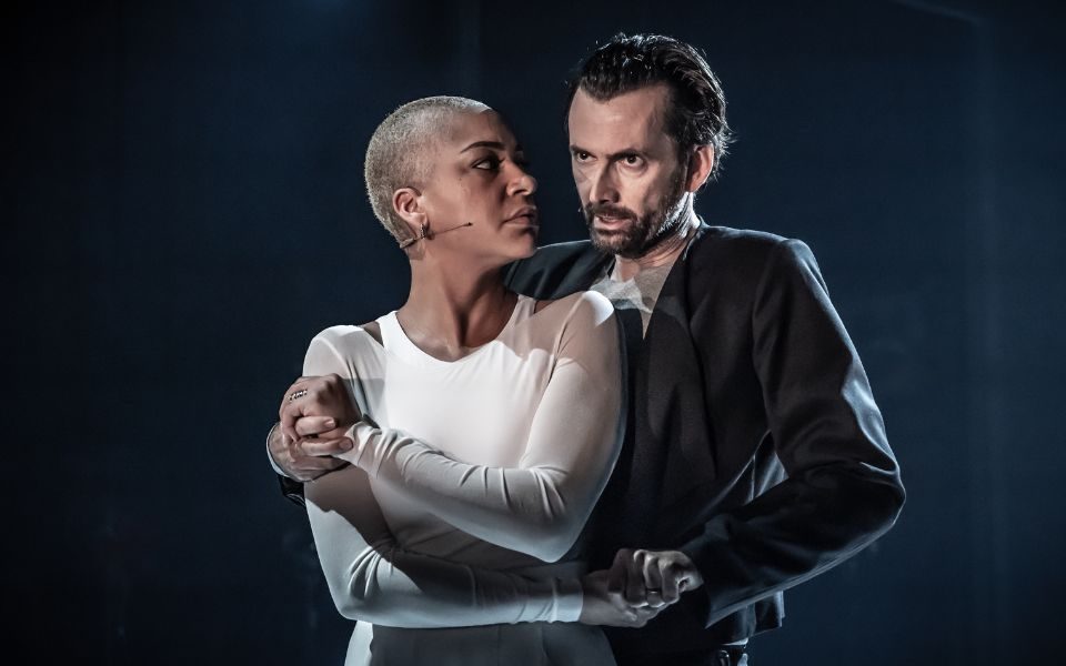 David Tennant as Macbeth in a suit jacket and grey t-shirt looking stern holding on to Cush Jumbo as Lady Macbeth wearing a white dress with shoulder cut outs who is looking back at David.