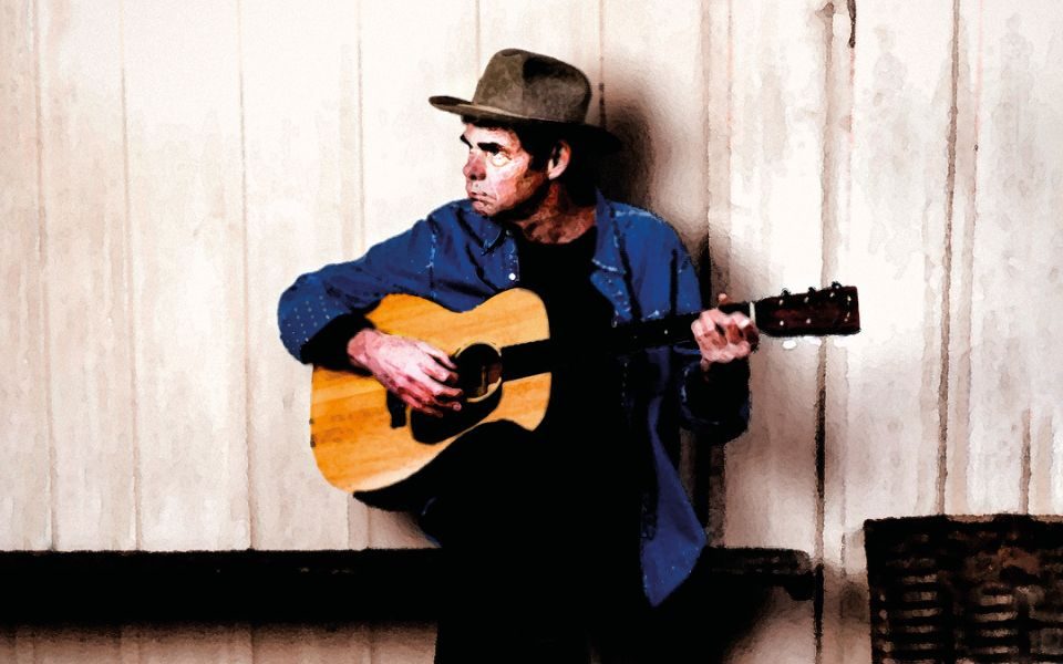 Rich Hall wearing a grey bowler hat playing against an acoustic guitar against a wooden wall.