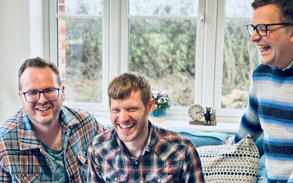 The three members of The Young Uns sat in patterned shirts laughing on a sofa.