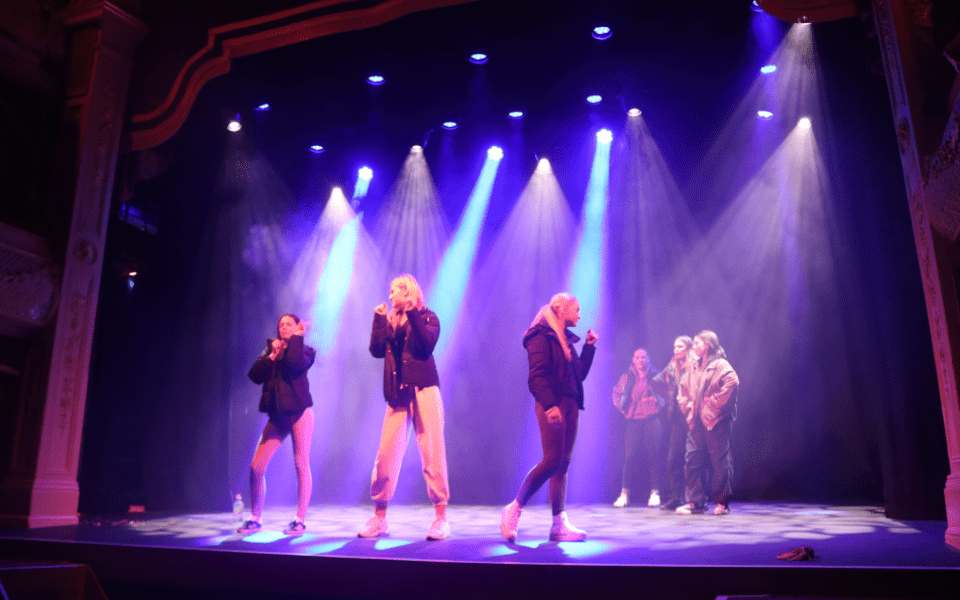 Students posing as if singing into microphones on stage at City Varieties with blue and purple lights on them.