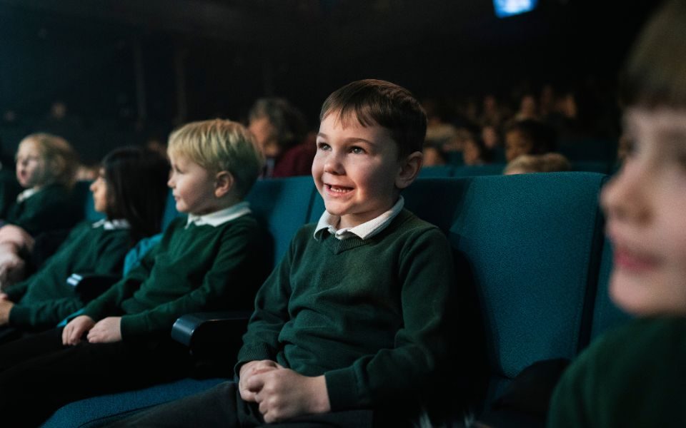 A row of primary schoolchildren in green uniform sit watching a live theatre performance and smiling