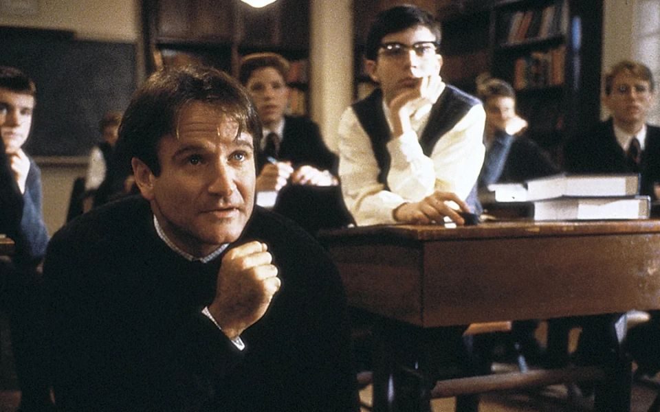 Robin Williams as a teacher in a classroom with boys lent on their desks behind him in school uniform.