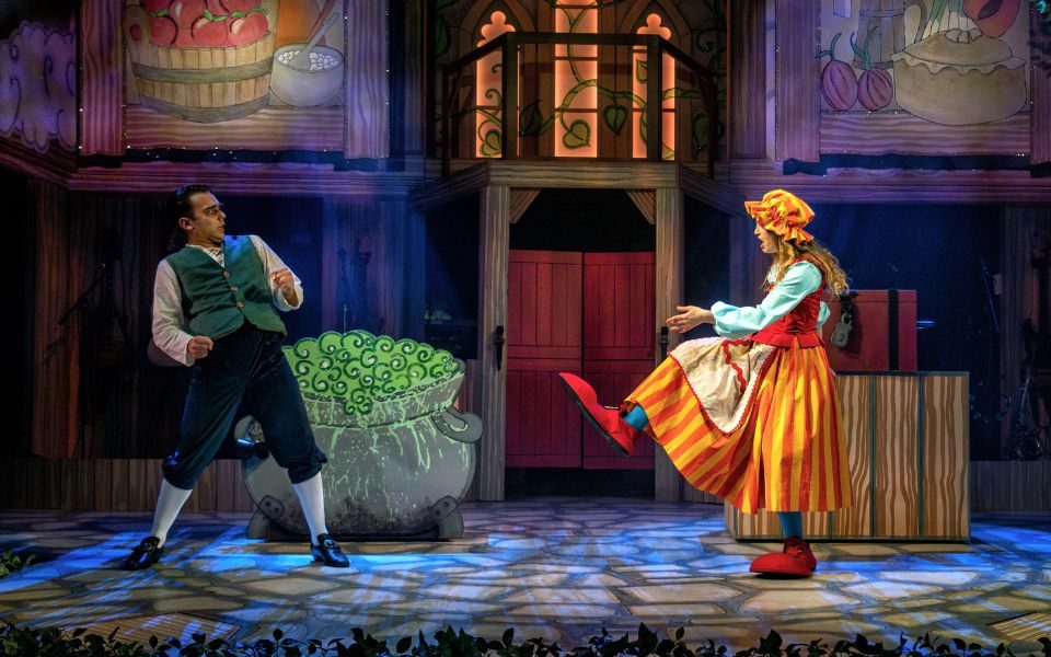 Stephanie Cremona as the Giant's wife in an orange dress showing off her big red clown shoes to Harry F Brown as Jack who looks aghast.