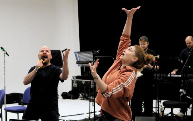 Kenny Davies sings dramatically into a microphone as he looks upwards. Next to him, Lucy Wells reaches upwards as if in a trance