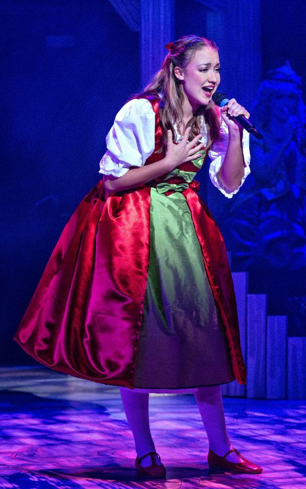 Lucy Ireland as Jill wearing a big red and green dress singing her heart out into a held microphone.
