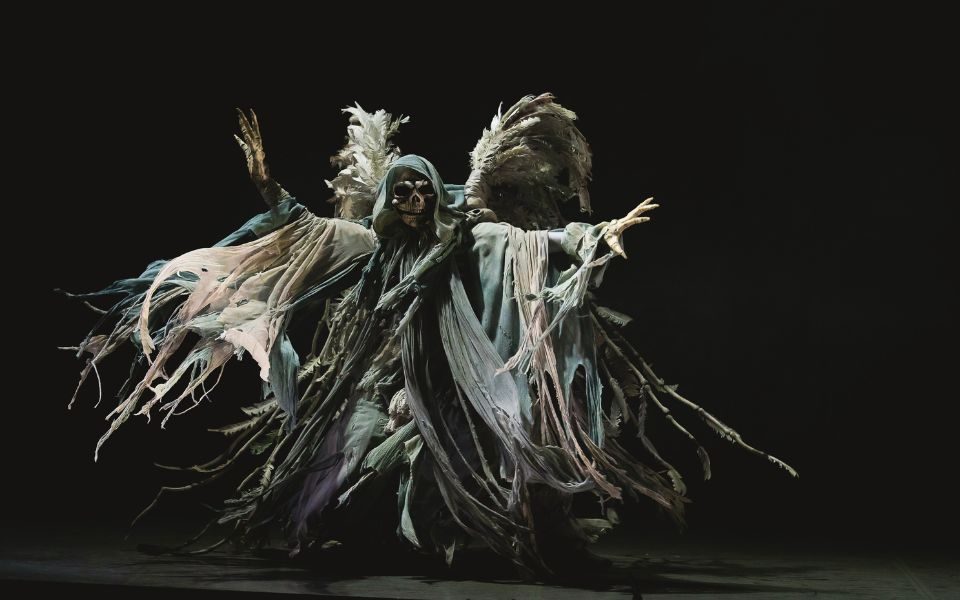 A dancer dressed in heavy layers of cloaks and a skull mask with large grey wings.