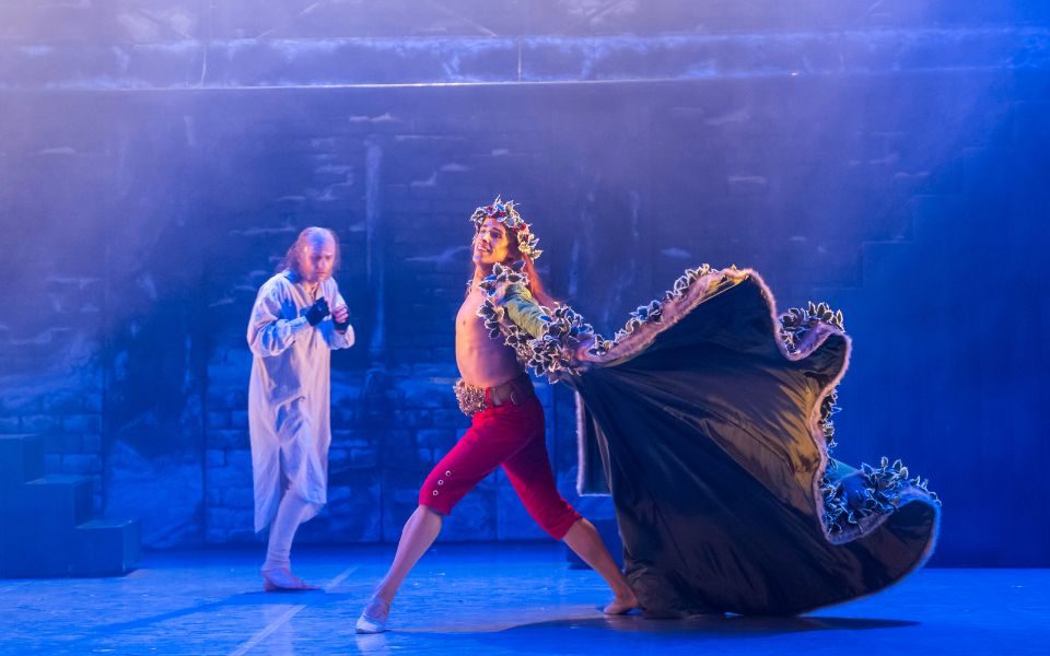 A dancer dressed in a old nightshirt as Scrooge stands watching a dancer dressed in red trousers and a long green cape adorned with ivy and greenery. The cape floats out dramatically as he walks across the stage.