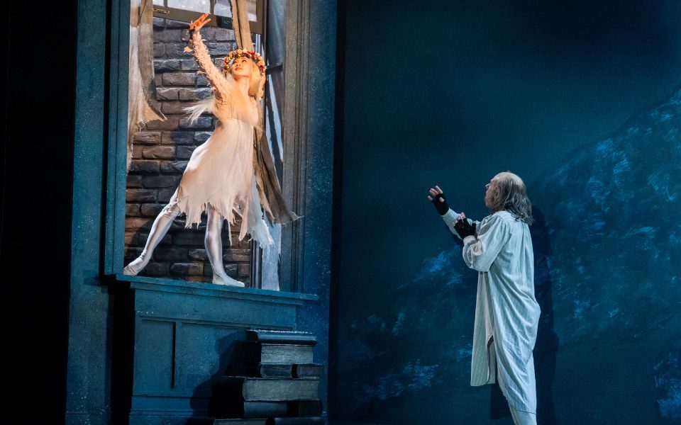 A dancer dressed in a floaty white dress and flower crown with flowing blonde hair stands on a window sill above a dancer dressed in a nightshirt as Scrooge.