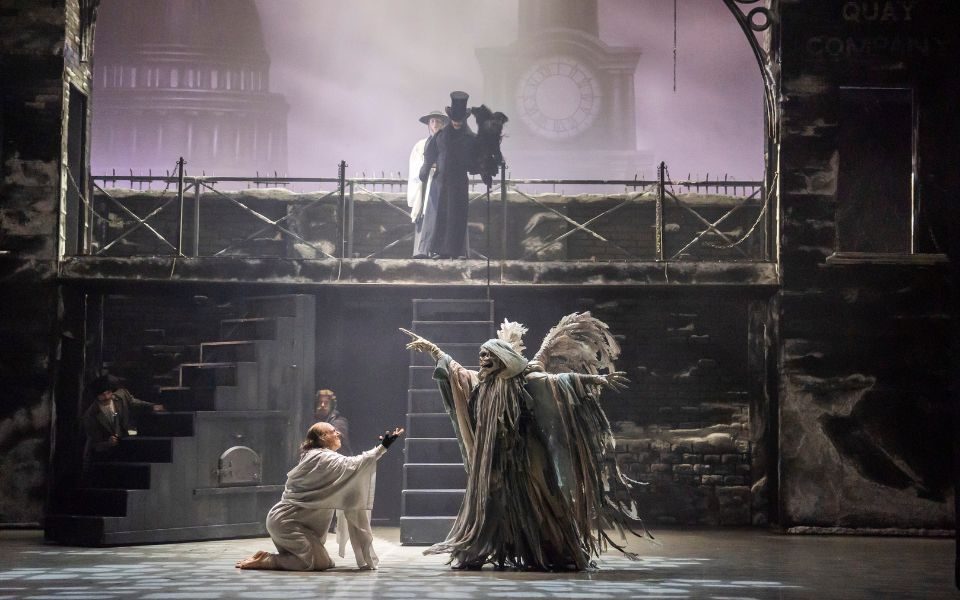 A dancer dressed in heavy layers of cloaks and a skull mask with large grey wings looms over a dancer dressed in a white nightgown as Scrooge. In the background a figure in a top hat looms on a raised platform.