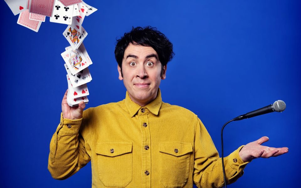 Pete Firman in a mustard-coloured shirt firing a pack of playing cards into the air with his right hand, his left levitating a cord microphone above it.