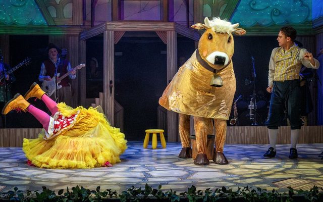 Panto cow Bessy on stage with panto Dame played by Simon Nock upside down on stage next to him with his underskirt covering his head and his legs in the air. Harry F Brown as Jack is looking at the cow.