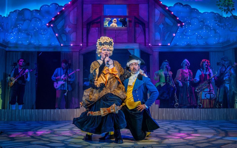Panto Dame Simon Nock in a big navy and gold dress sat on the knee of Dan Carter-Hope as the panto Squire. The cast as the band can be seen at the back of the stage.