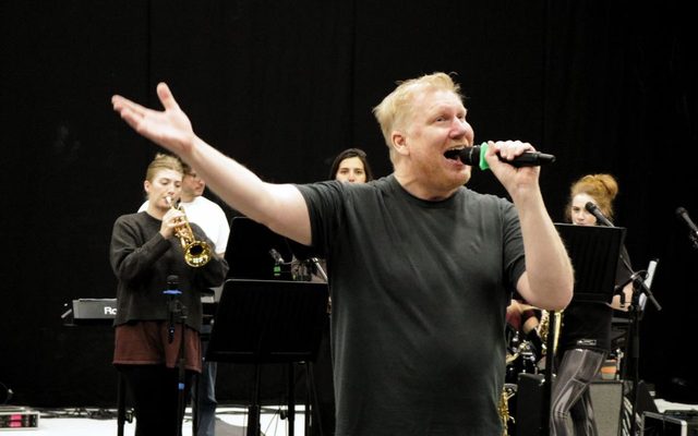 Simon Nock singing into a microphone in the rehearsal room, the band playing behind him