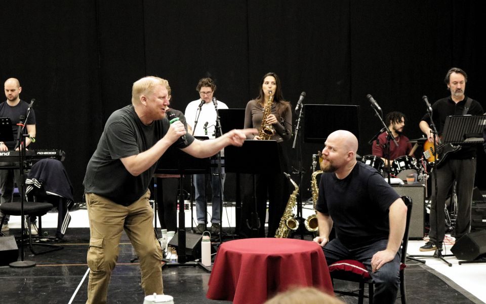 Simon Nock sings into a microphone as he wags his finger at Kenny Davies, who is sat at a small round table. The band plays behind them.