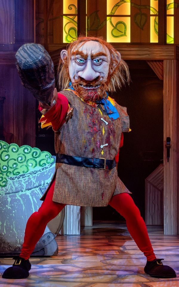 Panto giant with a big giant's mask on pointing a fake wooden club towards the camera. Fake blood and bones appear to be stuck to his beard and have fallen down his chest. He is wearing red tights and a brown tunic.