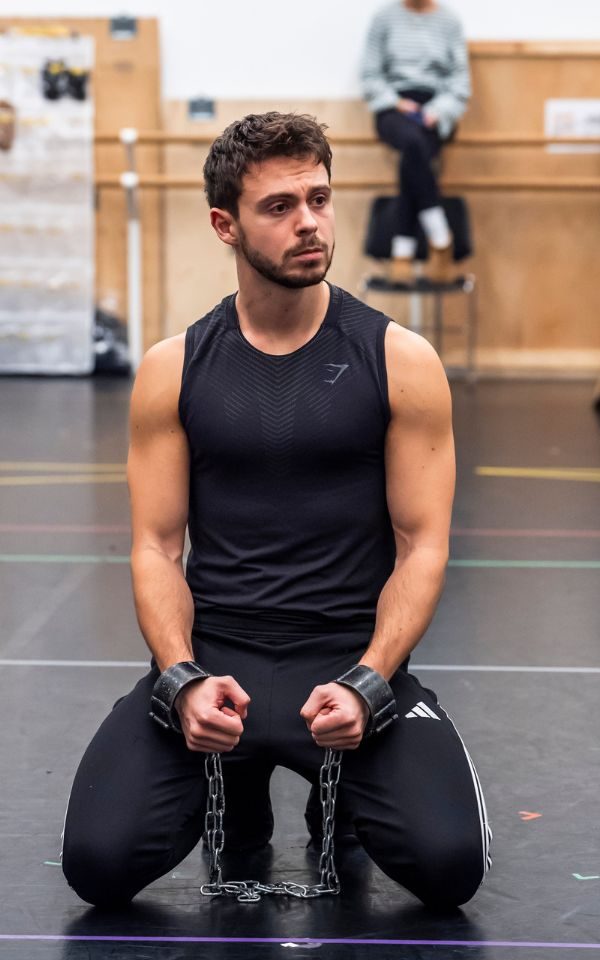 Adam Filipe with his hands handcuffed in chains kneeling down in rehearsals.