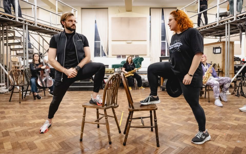 Vinny Coyle and Carrie Hope Fletcher face each other with one foot on a chair each, in rehearsals for Calamity Jane.