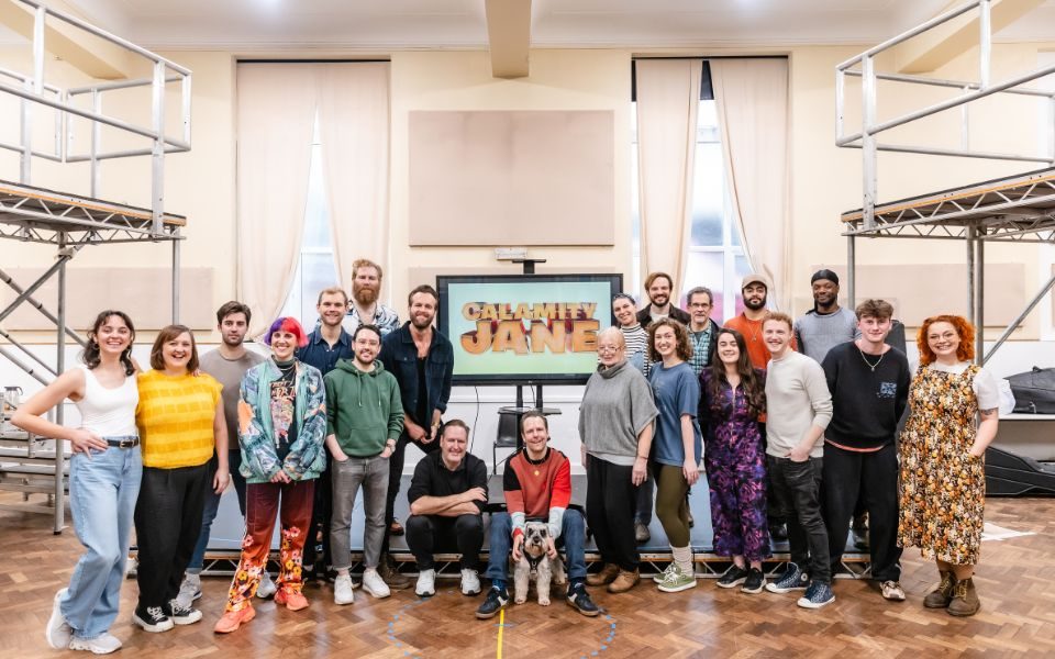 The cast of Calamity Jane grouped together with a poster for the show smiling at the camera in the rehearsal studio.