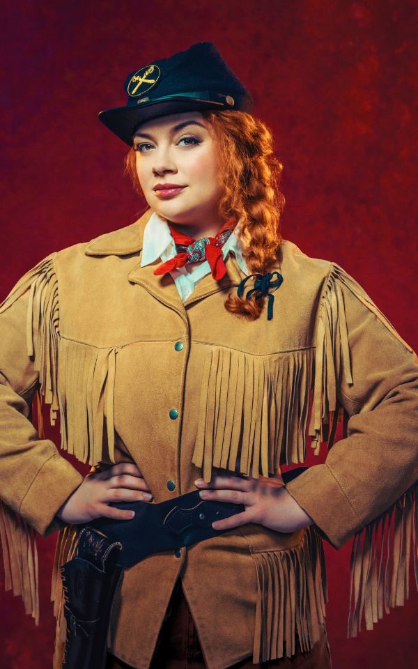 Carrie Hope Fletcher as Calamity Jane in a brown coat and black hat. She stands smirking with her hands on her hips.