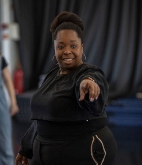 A drama facilitator in athletic wear stands and points towards the camera while smiling