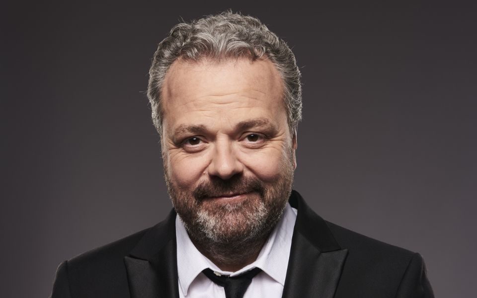 Hal Cruttenden smirking wearing a black suit with a think black tie.