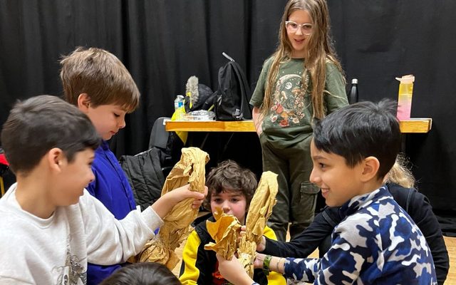 A group of young people at youth theatre huddle around crouching on the floor with paper puppets