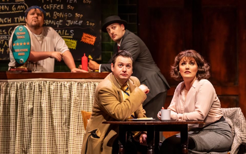 Richard J Hunt, Peter Watts, Sam Lupton and Georgina Hagen in Only Fools and Horses: The Musical. A man and woman sit at a table while a man in a suit leans on the bar towards the barman. They all stare out into the audience.