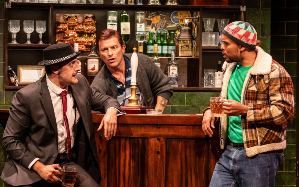 Three men talking at a bar in Only Fools and Horses: The Musical. One sits on a barstool, another holds a pint and the barman bends down.