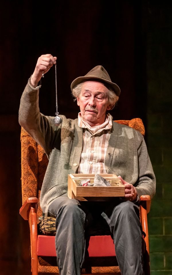 Paul Whitehouse in character sitting in an armchair and holding up a yoyo, wearing a brown hat and grey cardigan.