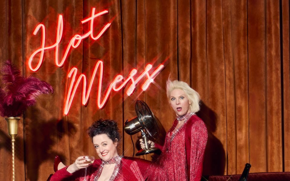 Helen and Ellie of Scummy Mummies in sparkly red outfits sat on a sofa a dtwo different levels with a bottle of bubbly on ice on the table next to them and a neon sign reading 'Hot Mess' behind them.