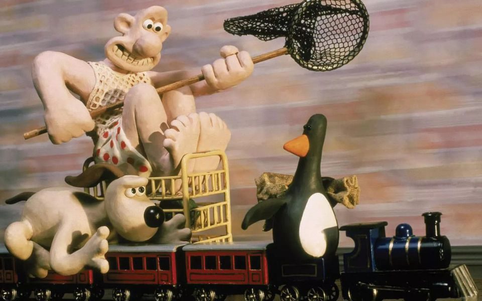 Wallace in his night vest and pyjama shorts sat on a trolley holding a fishing net on a pole while Gromit is trying to grab a suspicious-looking penguin riding on a toy train.