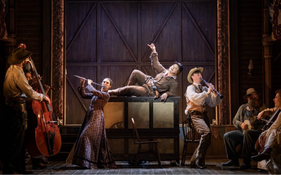 Carrie Hope Fletcher as Calamity Jane lying on top of a piano singing. The cast around are in costumes and playing instruments.