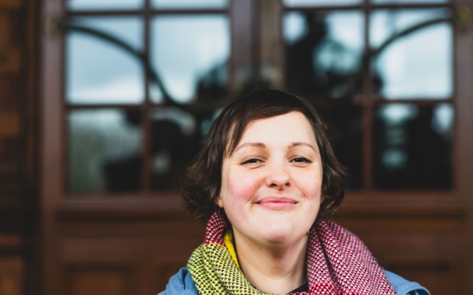 Josie Long wearing a lovely scarf smiling in front of a door with glass windows.