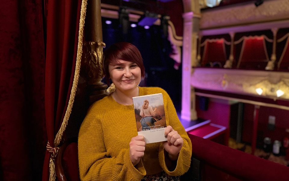 Imogen smiling with a copy of Little Women in the auditorium at City Varieties Music Hall