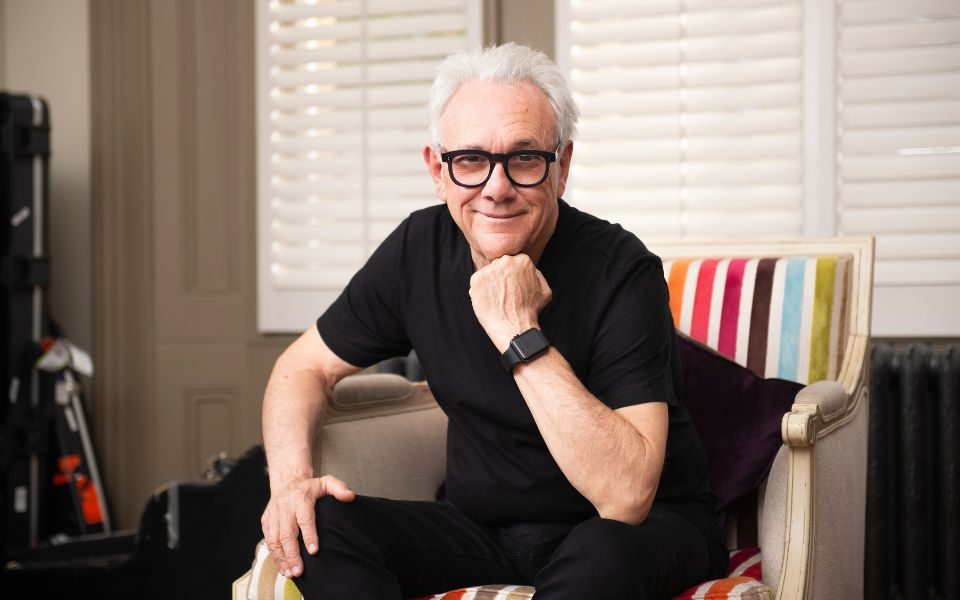 Trevor Horn in a black T-shirt sat on a rainbow-patterned chair, smiling at the camera and resting his chin on his hand.