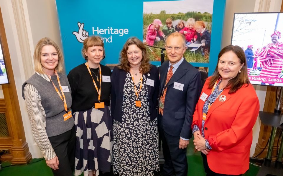 Five people, including Vicky Cheetham and Wendy Cook, smiling at the camera in front of a backdrop for Heritage Fund