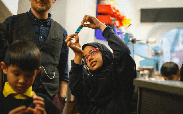 A young person with glasses and in a hijab is holding a piece of film reel up towards the lights to look through it.