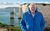 Ocean David Attenborough wearing a lovely warm blue jacket stood in front of impressive white cliffs that dart into a pale blue ocean.