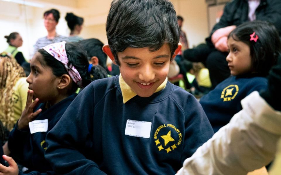 A primary schoolchild in uniform sits on the floor smiling and looking down, surrounded by other schoolchildren